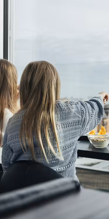 To personer sad indendørs og kiggede ud af et stort glasvindue med udsigt over bybilledet. Den ene peger udenfor, mens den anden observerer. En skål med snacks står på bordet foran dem. Begge har langt hår og bærer afslappet tøj.