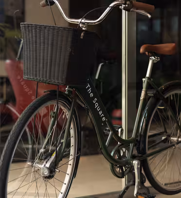 En grøn cykel i vintagestil med en sort vævet kurv er parkeret inde i en glasdør. Cyklen har brune lædergreb og et matchende sæde. Et stort grønt ornament er synligt i baggrunden indendørs.