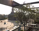 En solbelyst balkon med udsigt over et livligt bytorv. Et sparsomt træ i en plantekasse er i forgrunden, mens historiske murstensbygninger, inklusive en med et højt spir, er synlige i baggrunden under en delvist overskyet himmel.