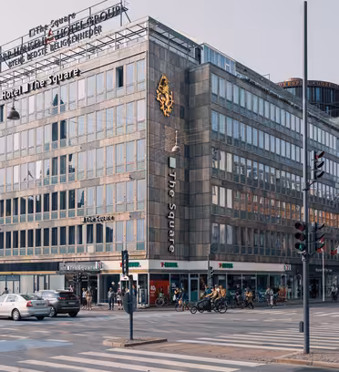 Travlt vejkryds i en by med en stor hjørnebygning med skiltning til "Hotel The Square". Cykler er parkeret udenfor, og fodgængere og biler er synlige. Arkitekturen omfatter rektangulære vinduer og urbane modernistiske designelementer.