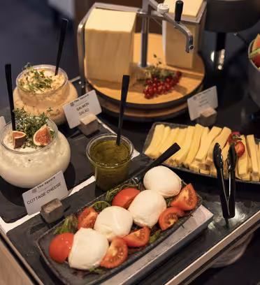 En buffet med mozzarella- og tomatsalat, hytteost, skiveskåret ost, vandmelonskiver og forskellige dips, pyntet med friske krydderurter og bær.