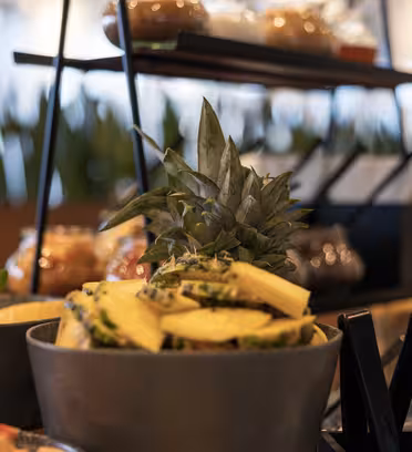 Nærbillede af en buffetopsætning med en skål skåret ananas med kronen som pynt. Friske mynteblade og diverse retter er i baggrunden, med en sløret udstilling af krukker og en række grønne planter, der tilføjer dybde.