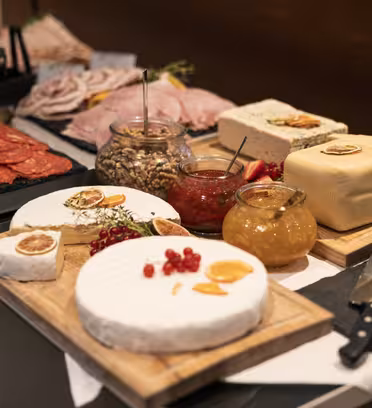 Et udvalg af forskellige oste, delikatessekød, nødder og frugter på træplader og bakker. Blandt de synlige ting kan nævnes runde oste, speget kød i skiver, krukker med nødder, marmelade og friske bær, pyntet med urter og tørrede citrusskiver.