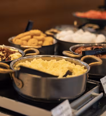 Et buffetbord med diverse morgenmadsretter i metalgryder. Synlige ting omfatter røræg, baconstykker, pølser, donuts, kogte æg og meget mere. Hver gryde har en serveringsske, og små skilte mærker retterne.