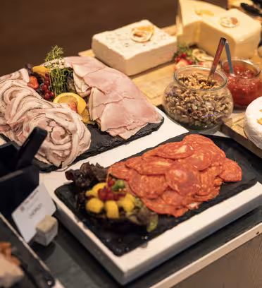 En buffet med forskellige kødstykker, oste og pynt. Tallerkener viser rullet skinke, salami og paté sammen med krukker med krydderier. Ostblokke ledsages af garniture som tørret citrus og urter på en træbord.