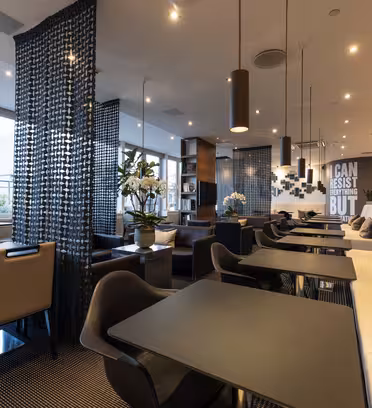 Et moderne caféinteriør med sorte borde og en blanding af beige og sorte stole. Dekorative paneler deler rummet op, og store vinduer giver naturligt lys. En grå sofa med hynder langs den ene væg, under et citat med fed tekst.