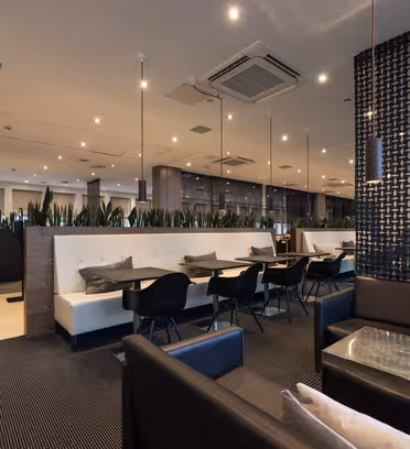 Moderne restaurantinteriør med et slankt, moderne design. Har hvide båse, sorte stole og potteplanter som skillevægge. En mønstret sort væg tilføjer kontrast, og overheadbelysning skaber en varm stemning.