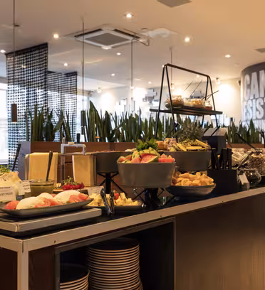 En moderne buffetopstilling med en række forskellige retter vist på en disk, herunder salater, frugter og desserter. Baggrunden byder på høje planter og et stærkt oplyst spiseområde. Indretningen er slank med et moderne design.