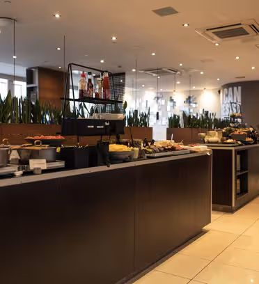 En rummelig, moderne buffet har forskellige retter på diskene, herunder kager og frugter. Baggrunden viser indendørs planter og spejle, mens blød belysning forstærker den moderne stemning.