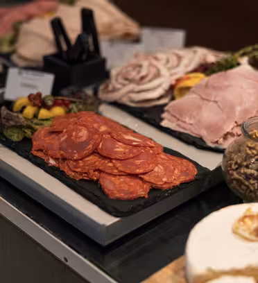 Et fad med skåret speget kød, inklusive pepperoni og skinke, vises på en buffet. Ved siden af er en skål med valnødder, et ostehjul og diverse garniture synlige. Andet pålæg står i baggrunden på sorte serveringsbakker.