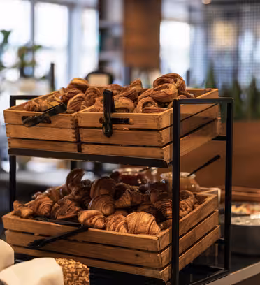 Trækasser fyldt med croissanter og kager er udstillet på en metalstativ. I baggrunden ses et sløret billede af et moderne caféinteriør med nogle grønne planter og et par lånere.