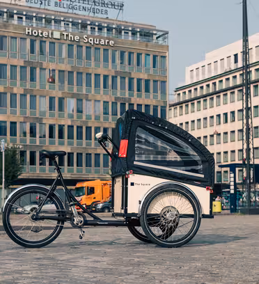 En trehjulet ladcykel med overdækket siddeområde er parkeret på en brostensbelagt gade. Bag det er flere etagers bygninger, herunder et hotel ved navn "The Square." Scenen er urban med en klar himmel.