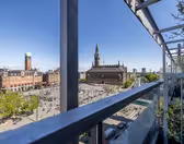 Udsigt fra en balkon med udsigt over en stor byplads med gående mennesker, omgivet af historiske bygninger og et højt klokketårn, under en klar blå himmel.