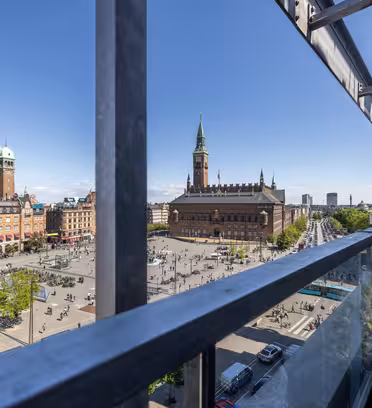 Udsigt fra en balkon med udsigt over en stor byplads med gående mennesker, omgivet af historiske bygninger og et højt klokketårn, under en klar blå himmel.