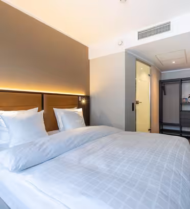Et moderne hotelværelse med en stor, pænt redt seng, hvidt sengetøj, to puder, dæmpet belysning, en åben garderobe, et minikøleskab og et badeværelse med glasvægge i baggrunden.