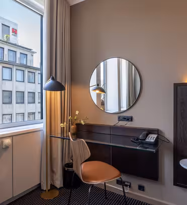 Et moderne hotelværelseshjørne med et glasskrivebord, stol, rundt spejl, skrivebordslampe og telefon; en seng er delvist synlig, og store vinduer lukker naturligt lys ind og viser bygninger udenfor.