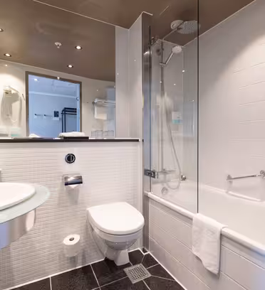 Moderne badeværelse med hvide fliser, en glasvask, væghængt toilet, badekar med bruser, stort spejl, håndklædeholder og klar loftsbelysning. Et hvidt håndklæde hænger på badekarskanten.