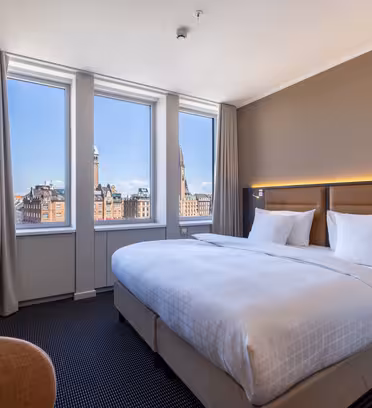Et moderne hotelværelse med en stor, pænt redt seng, tre høje vinduer med udsigt over byen, beige vægge, blød belysning og møbler i neutrale farver.