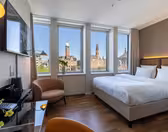 Et moderne hotelværelse med en stor seng, et tv, en brun lænestol, et skrivebord og tre store vinduer med udsigt over byen med historiske bygninger og tårne under en klar blå himmel.