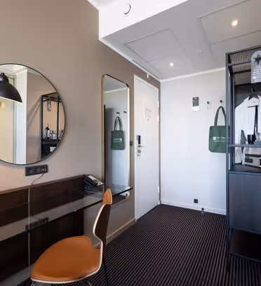 Moderne hotelværelse med en brun stol ved et glasskrivebord, rundt spejl, væglampe og et klædeskab med hylder og hængende ting. Et stort vægspejl og en grøn mulepose hænger ved den hvide dør.