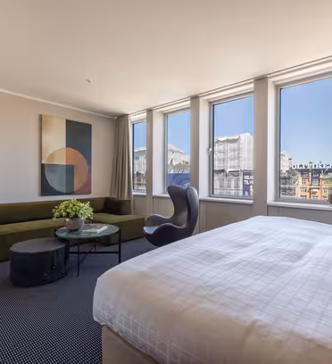 Et moderne hotelværelse med en stor seng, grøn sofa, rundt sofabord og lænestol, gulv-til-loft-vinduer med udsigt over byen og naturligt lys samt abstrakt kunst på væggen.