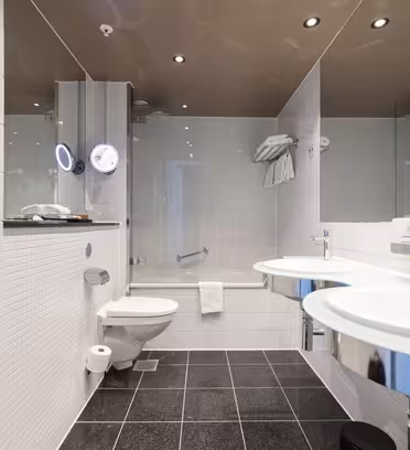 Moderne hotelbadeværelse med hvide flisebelagte vægge, sort flisebelagt gulv, to runde glasvaske med spejle, et vægmonteret toilet, håndklædestativer og et badekar med glasvæg og monteret brusehoved.