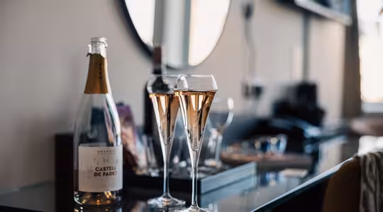 To glas rosé mousserende vin står på et glasbord ved siden af en åben flaske mærket Castell de Fades med slørede genstande og et spejl i baggrunden.