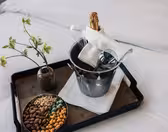 På en bakke på en seng står en vase med grønne blomster, en skål med forskellige nødder og en metalisspand med en champagneflaske, to glas og en foldet hvid serviet.