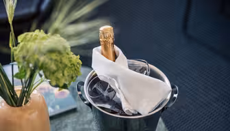 En kold flaske champagne pakket ind i en hvid serviet står i en isspand ved siden af to glas, med en vase med grønne blomster i nærheden på et bord.