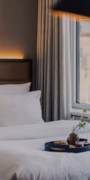 Et moderne, hyggeligt hotelværelse med pænt redte enkeltsenge, hvidt sengetøj, et stort vindue med beige gardiner og en bakke med morgenmadsprodukter på sengen. En vase med blomster står i vindueskarmen.