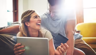 Et smilende par sidder sammen i et soloplyst rum og kigger på en tablet. Kvinden holder apparatet og et kreditkort, mens manden læner sig over hendes skulder. En sofa og en gul stol er i baggrunden.