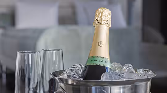 En flaske Brut Nature champagne køler i en isfyldt metalspand, ledsaget af to tomme champagnefløjter. En sløret seng med hvide puder er synlig i baggrunden, hvilket tyder på hyggelige omgivelser.