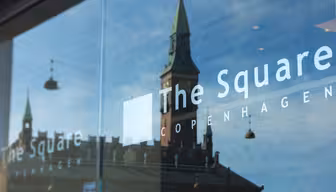 Refleksion af en gammel bygning med et grønt spir i glasvinduet på "The Square" hotel i København. Refleksionen viser indviklede arkitektoniske detaljer af bygningen under en klar himmel.