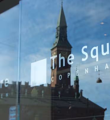 Refleksion af en gammel bygning med et grønt spir i glasvinduet på "The Square" hotel i København. Refleksionen viser indviklede arkitektoniske detaljer af bygningen under en klar himmel.