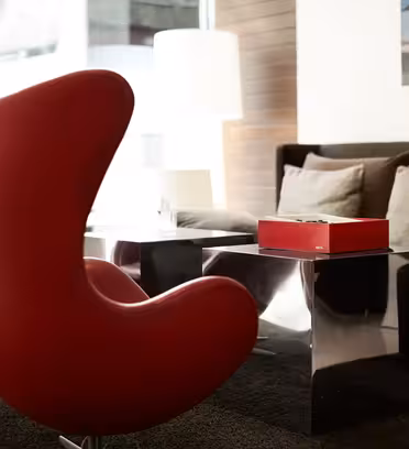 Moderne stue med to røde, buede lænestole ud mod et glas sofabord. En brun sofa med beige hynder er i baggrunden, sammen med en bordlampe og vægkunst. Rummet er godt oplyst af naturligt lys.