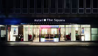 Gadebillede af en hotelindgang ved navn "Hotel The Square" om natten. Bygningen har store vinduer, der viser en veloplyst lobby med moderne møbler, kunstværker og potteplanter. Det oplyste skilt tilføjer et indbydende touch.
