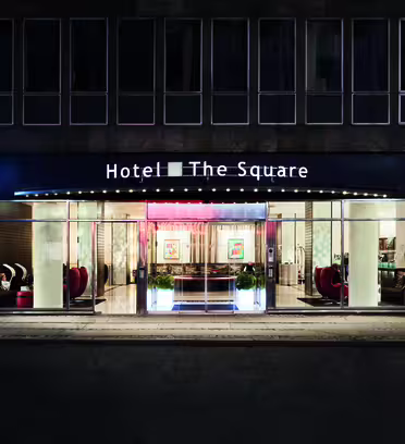 Gadebillede af en hotelindgang ved navn "Hotel The Square" om natten. Bygningen har store vinduer, der viser en veloplyst lobby med moderne møbler, kunstværker og potteplanter. Det oplyste skilt tilføjer et indbydende touch.