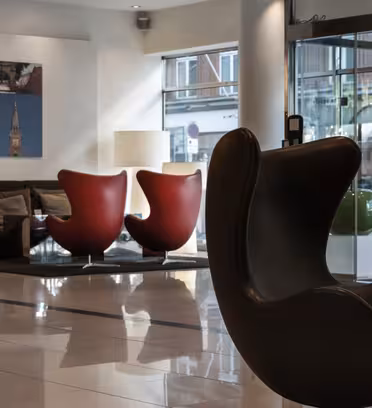 Moderne hotelobby med blankt flisegulv, med en mørk moderne stol i forgrunden, to røde æggestole nær et vindue og en sofa med puder. Vægkunst med bybilleder hænger på en hvid væg, og glasdøre afslører en bygade udenfor.