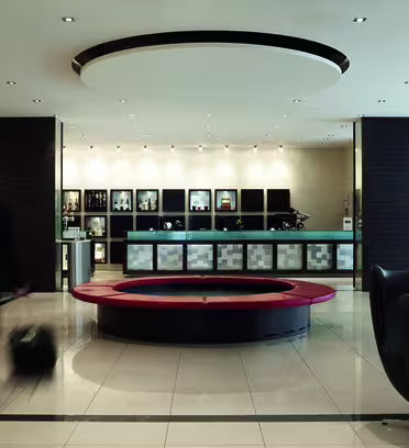 En rummelig hotellobby med et moderne design. En sløret mand i jakkesæt går forbi med en rullekuffert. Receptionen er i centrum med et cirkulært rødt og sort siddeområde foran. Minimalistiske stole er placeret rundt i lobbyen.