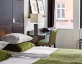 Et moderne hotelværelse med en pæn opredt seng med grønt og hvidt sengetøj, en sengegavl i træ og et glasskrivebord med lampe og telefon. Indrammet kunst er på væggen, og to store vinduer giver naturligt lys og udsigt udenfor.
