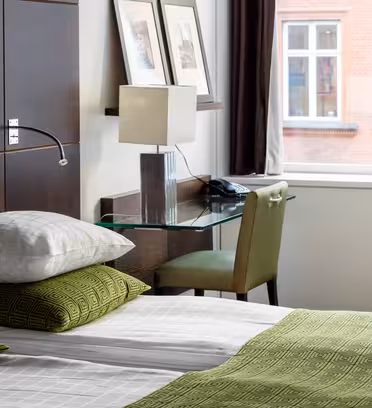 Et moderne hotelværelse med en pæn opredt seng med grønt og hvidt sengetøj, en sengegavl i træ og et glasskrivebord med lampe og telefon. Indrammet kunst er på væggen, og to store vinduer giver naturligt lys og udsigt udenfor.