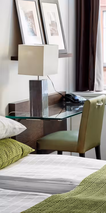 Et moderne hotelværelse med en pæn opredt seng med grønt og hvidt sengetøj, en sengegavl i træ og et glasskrivebord med lampe og telefon. Indrammet kunst er på væggen, og to store vinduer giver naturligt lys og udsigt udenfor.