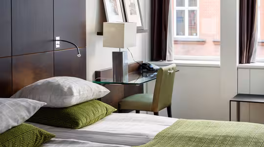 Et moderne hotelværelse med en pæn opredt seng med grønt og hvidt sengetøj, en sengegavl i træ og et glasskrivebord med lampe og telefon. Indrammet kunst er på væggen, og to store vinduer giver naturligt lys og udsigt udenfor.