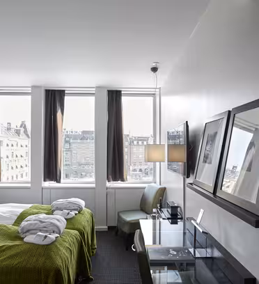 Moderne hotelværelse med to enkeltsenge og grønt sengetøj. Værelset har skrivebord, stol og vægmonteret tv. Store vinduer med brune gardiner tilbyder udsigt over byen. To indrammede billeder er vist på en hylde over skrivebordet.