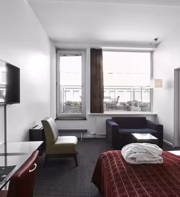 Moderne hotelværelse med en dobbeltseng med rødt sengetøj, en foldet hvid morgenkåbe på toppen og et lille loungeområde. Der er et fladskærms-tv på væggen, et skrivebord med en stol og store vinduer, der tillader naturligt lys.