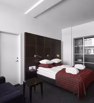 Et moderne hotelværelse med en dobbeltseng dækket af rødt og hvidt sengetøj. Der er to morgenkåber på sengen, en sort sofa, et bord med glasplade og et klædeskab. Værelset har et minimalistisk design med mørkt tæppe og sengegavl i træ.