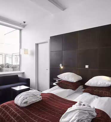 Et moderne hotelværelse har to enkeltsenge med rødmønstrede sengetæpper og hvide puder. En sort sofa og et bord med glasplade sidder ved et stort vindue med udsigt over en balkon. Rummet har mørkt træpanel og minimalistisk indretning.