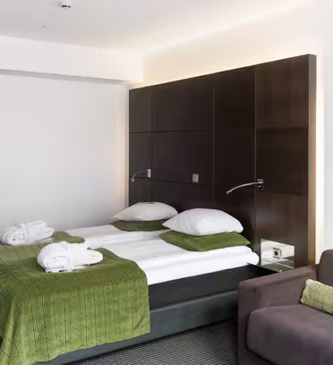 Moderne hotelværelse med dobbeltseng prydet med grønt sengetøj og puder. To hvide klæder er placeret på sengen. Der er en mørk sofa med en grøn pude og blød belysning over sengens hovedgærde i træ. Gulvet er gulvtæppe i mørkegrå.