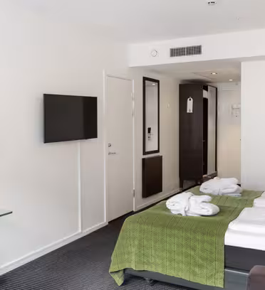 Hotelværelse med en pæn lavet dobbeltseng med grønt sengetæppe og foldede håndklæder. Et fladskærms-tv er monteret på væggen overfor et glasbord med en lampe. Der er et klædeskab, en lille skraldespand og et spejl i fuld længde nær indgangen.