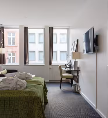 Et hotelværelse med to pænt opredte senge med grønne sengetæpper og hvide morgenkåber. Et skrivebord med en stol og et fladskærms-tv står op ad væggen. Store vinduer giver udsigt til tilstødende bygninger.